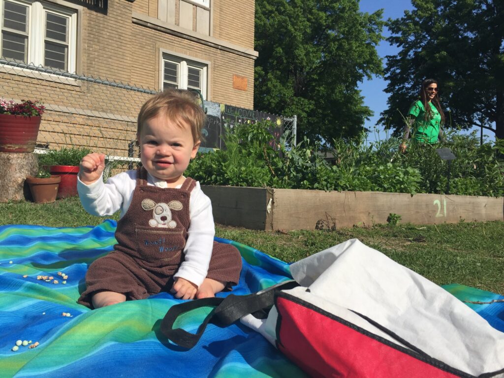 This infant comes with his mom at Mallinckrodt school garden and brings his own seating. His mom weeds and plants while he sits and waves at everyone!